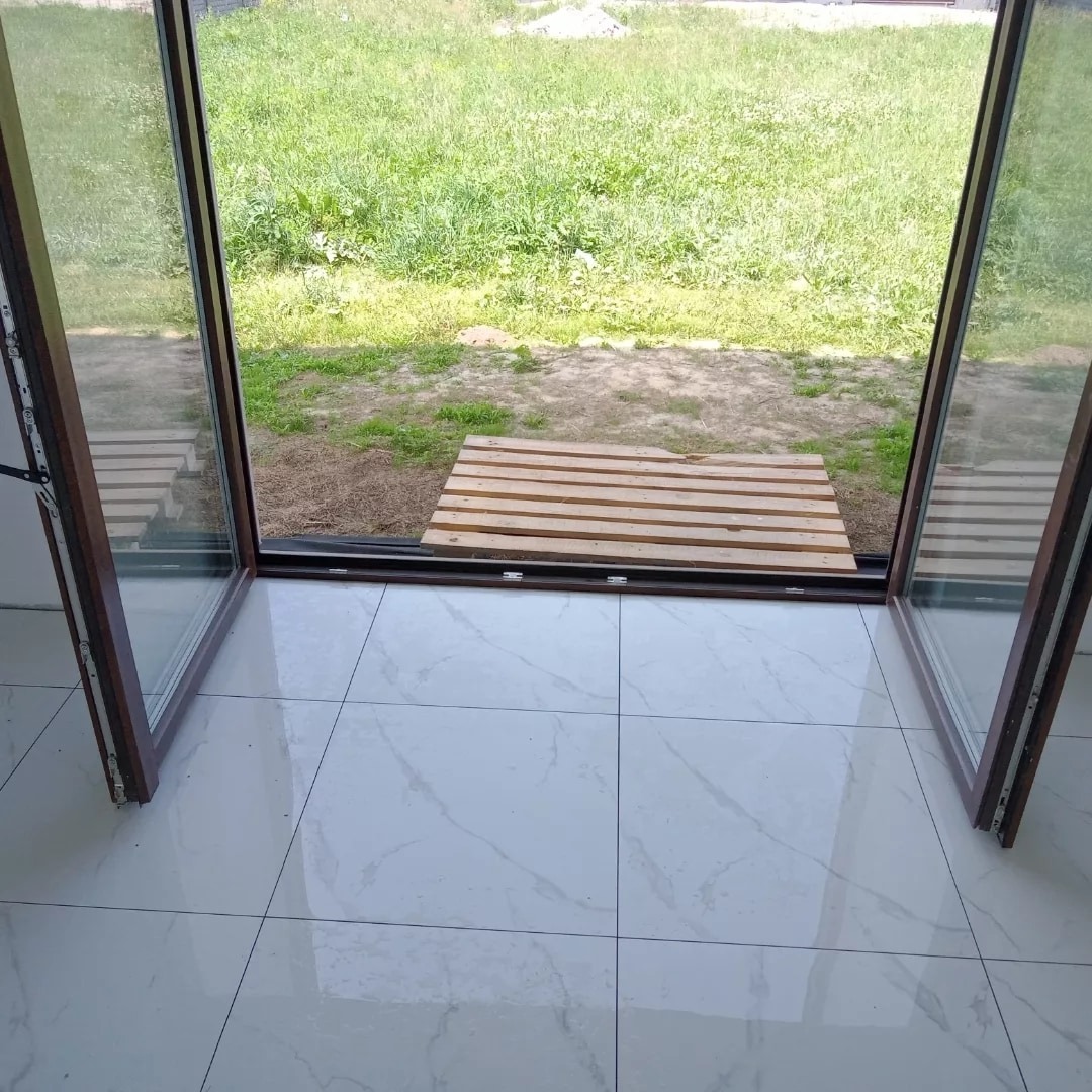 Widok z wnętrza na zewnątrz przez otwarte, brązowe drzwi balkonowe. Na podłodze wewnątrz białe płytki imitujące marmur. Na zewnątrz trawa i drewniana paleta.