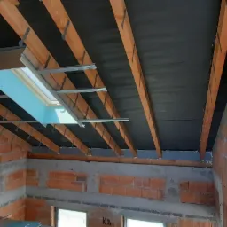Poddasze w trakcie ocieplania z widocznym oknem dachowym w metalowej ramie, czarną membraną na krokwiach i czerwoną cegłą na ścianach.