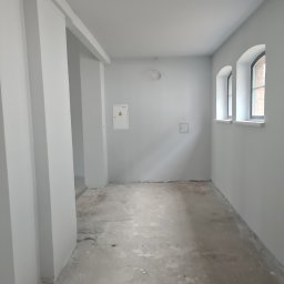 Od Nowa - Wnętrze w trakcie przygotowania do prac wykończeniowych, świeżo pomalowane ściany na szaro, surowa betonowa podłoga, dwa łukowe okna w tle.