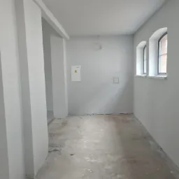 Wnętrze w trakcie przygotowania do prac wykończeniowych, świeżo pomalowane ściany na szaro, surowa betonowa podłoga, dwa łukowe okna w tle.