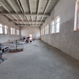 Od Nowa - Przestronne wnętrze w trakcie przygotowania ścian pod malowanie, widoczne narzędzia, rusztowania i szpachlowane ściany w dużej hali z rzędem okien i drewnianym stropem.