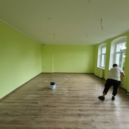 Od Nowa - Jasnozielone ściany świeżo pomalowanego pokoju z drewnopodobnymi panelami podłogowymi; widoczne wystające kable ze sufitu i pracownik malujący dolną część ściany przy oknie.