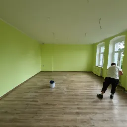 Jasnozielone ściany świeżo pomalowanego pokoju z drewnopodobnymi panelami podłogowymi; widoczne wystające kable ze sufitu i pracownik malujący dolną część ściany przy oknie.