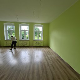 Od Nowa - Malowanie ścian na zielono w pokoju z drewnianą podłogą, widoczne trzy okna z białymi ramami i rysunki na grzejnikach, mężczyzna zamiata podłogę po malowaniu.