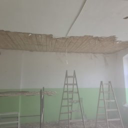 Od Nowa - Demontaż starego sufitu w pomieszczeniu z zielonym pasem na ścianie; widoczne drewniane listwy i fragmenty tynku, dwie drabiny ustawione w pokoju, okno z parapetem również pokrytym resztkami...