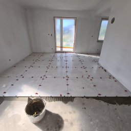 Od Nowa - Jasnoszare płytki podłogowe w trakcie układania, z czerwonymi klipsami poziomującymi, widoczny fragment nałożonego kleju, białe ściany w surowym stanie, wiadro z zaprawą w rogu.