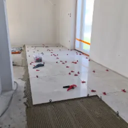 Układanie jasnych płytek podłogowych z użyciem systemu poziomowania, widoczny klej z charakterystycznymi rowkami, czerwone klipsy i kliny, w tle okno zabezpieczone folią.