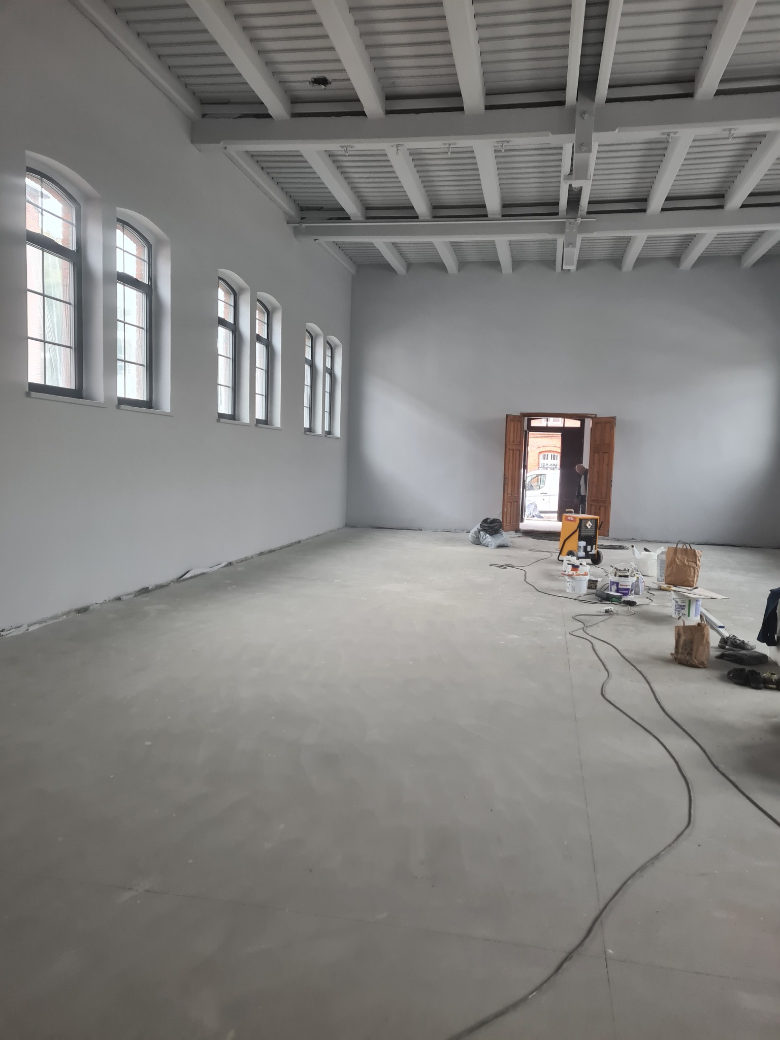 Duże, jasne pomieszczenie po świeżym malowaniu na szaro, z charakterystycznymi łukowymi oknami, drewnianą konstrukcją sufitu i sprzętem malarskim w tle.