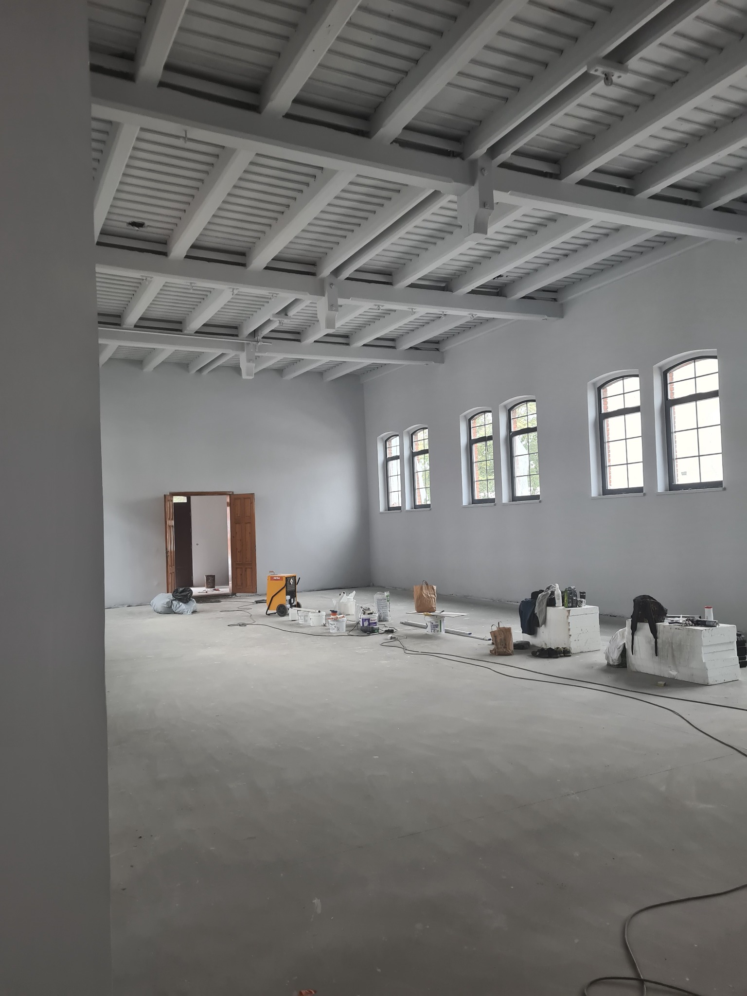 Duże, jasne pomieszczenie w trakcie malowania na szaro, widoczne narzędzia malarskie, drabina, otwarte drzwi do sąsiedniego pokoju i charakterystyczny, kratowany sufit.