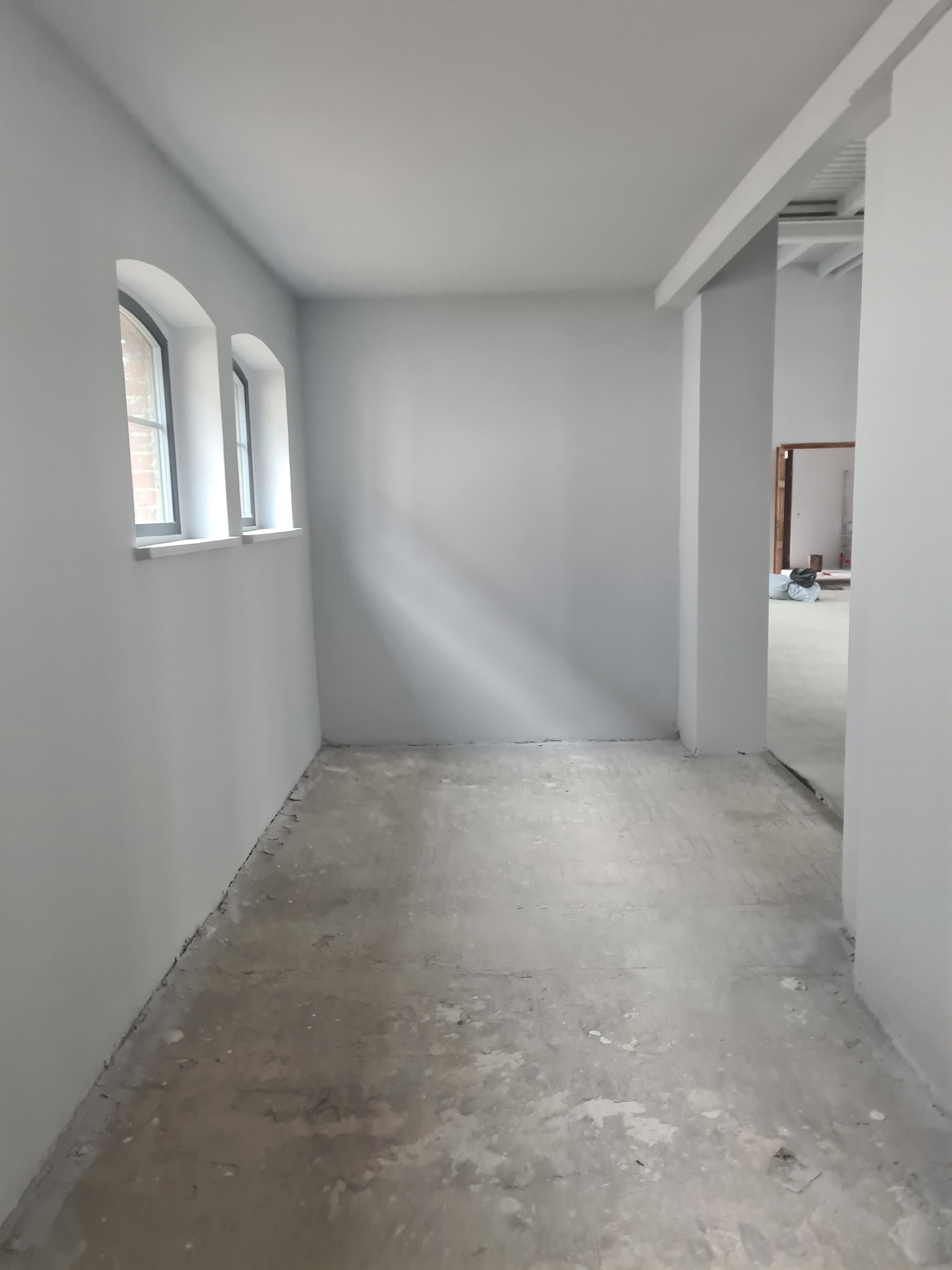Wnętrze korytarza po malowaniu na szaro, z widocznymi trzema łukowatymi oknami i surową podłogą przygotowaną do dalszych prac wykończeniowych.