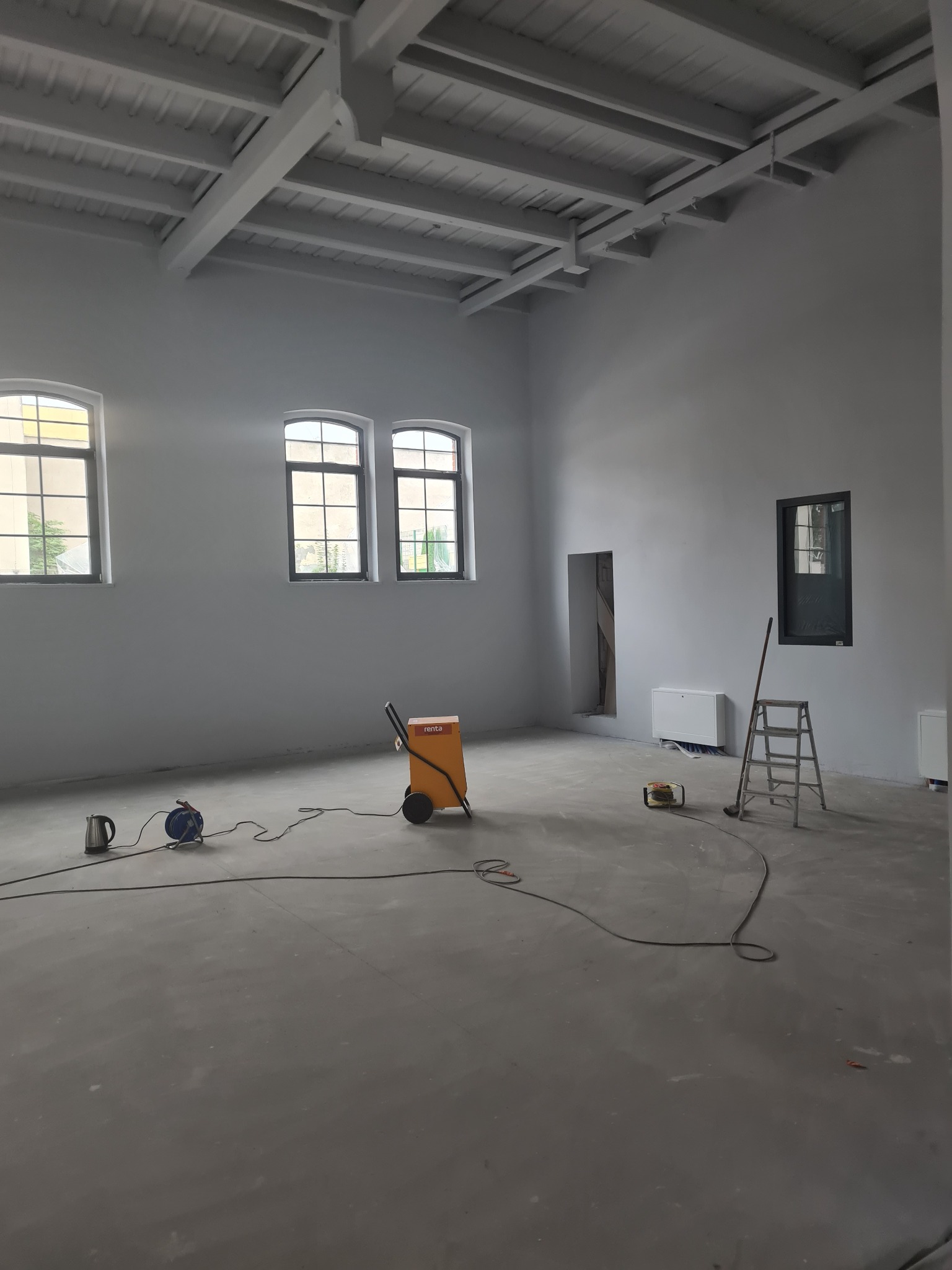 Duże, pomalowane na szaro pomieszczenie z trzema oknami po lewej stronie, osuszaczem powietrza Renta na kółkach, przedłużaczem, czajnikiem elektrycznym, grzejnikiem, drabiną i szczotką przy ścianie...