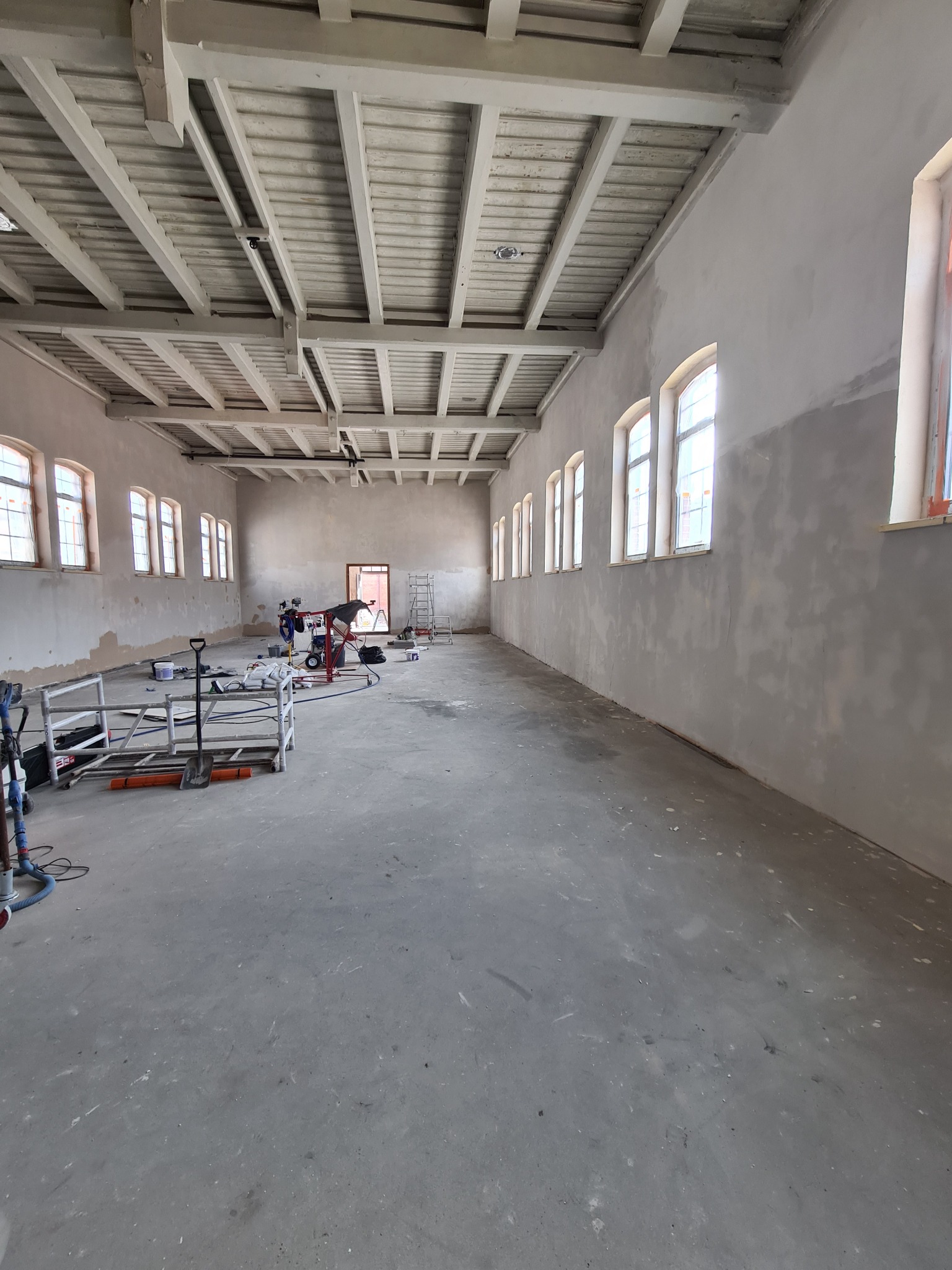 Przestronne wnętrze w trakcie przygotowania ścian pod malowanie, widoczne narzędzia, rusztowania i szpachlowane ściany w dużej hali z rzędem okien i drewnianym stropem.