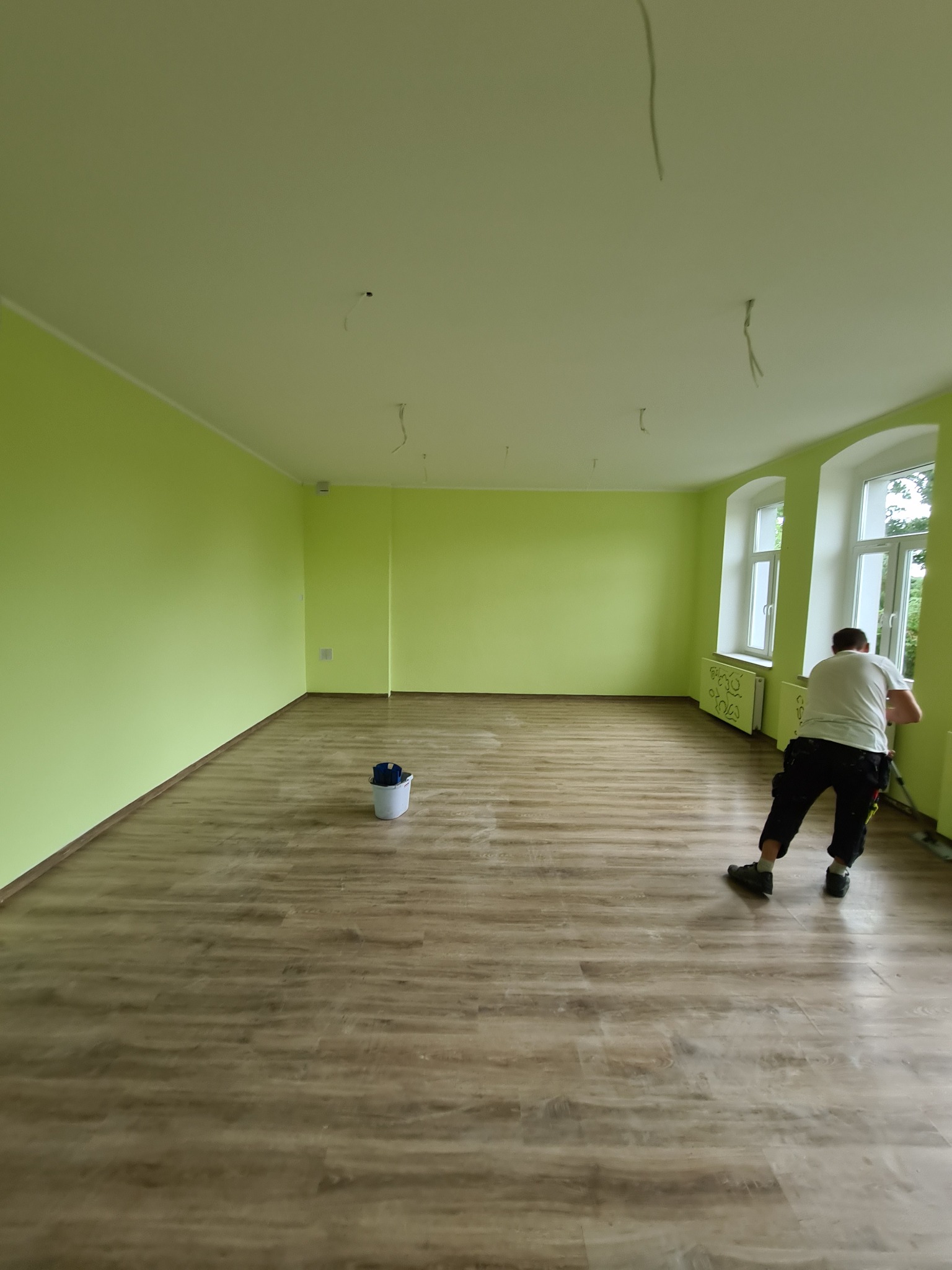 Jasnozielone ściany świeżo pomalowanego pokoju z drewnopodobnymi panelami podłogowymi; widoczne wystające kable ze sufitu i pracownik malujący dolną część ściany przy oknie.