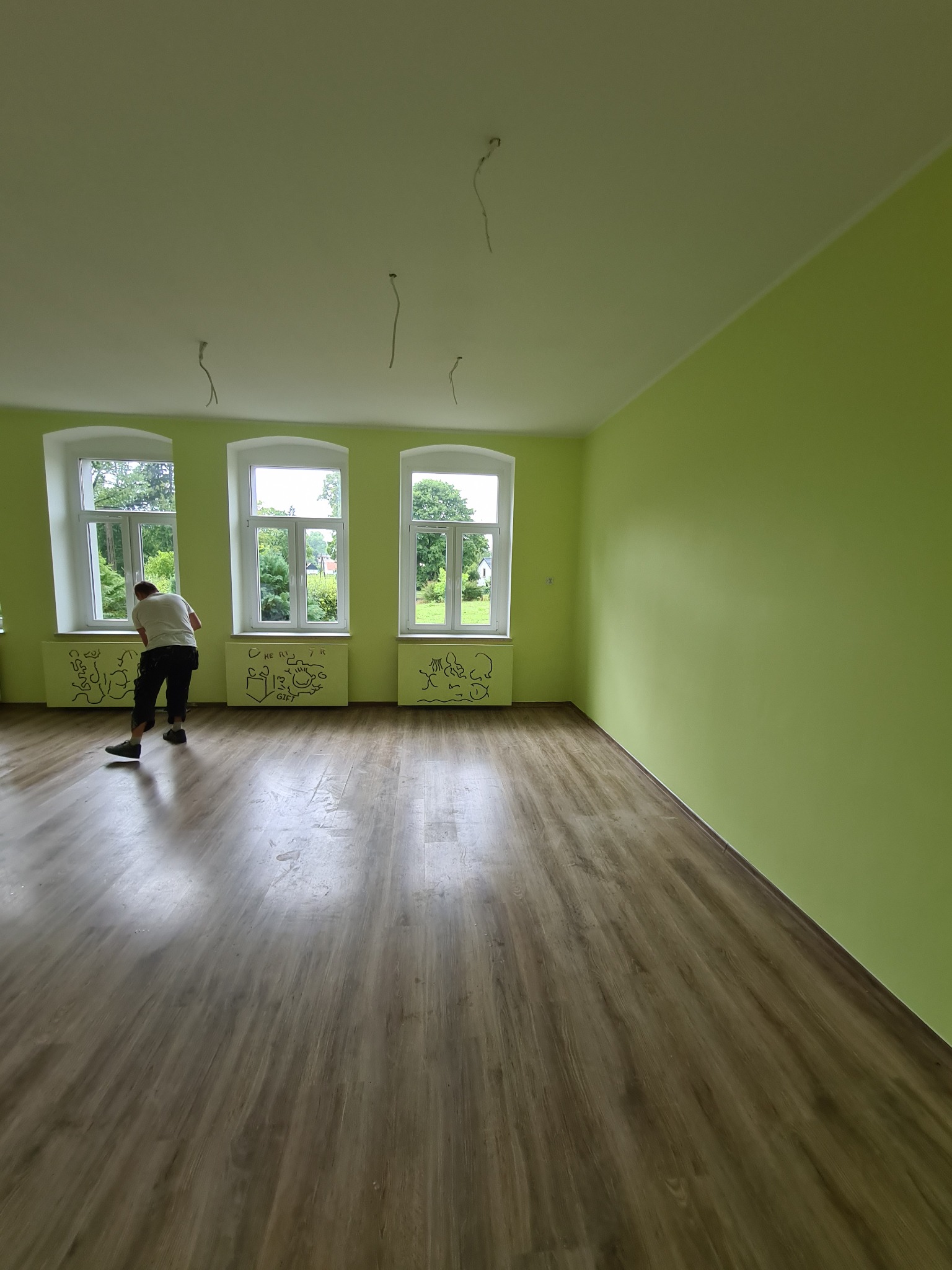 Malowanie ścian na zielono w pokoju z drewnianą podłogą, widoczne trzy okna z białymi ramami i rysunki na grzejnikach, mężczyzna zamiata podłogę po malowaniu.