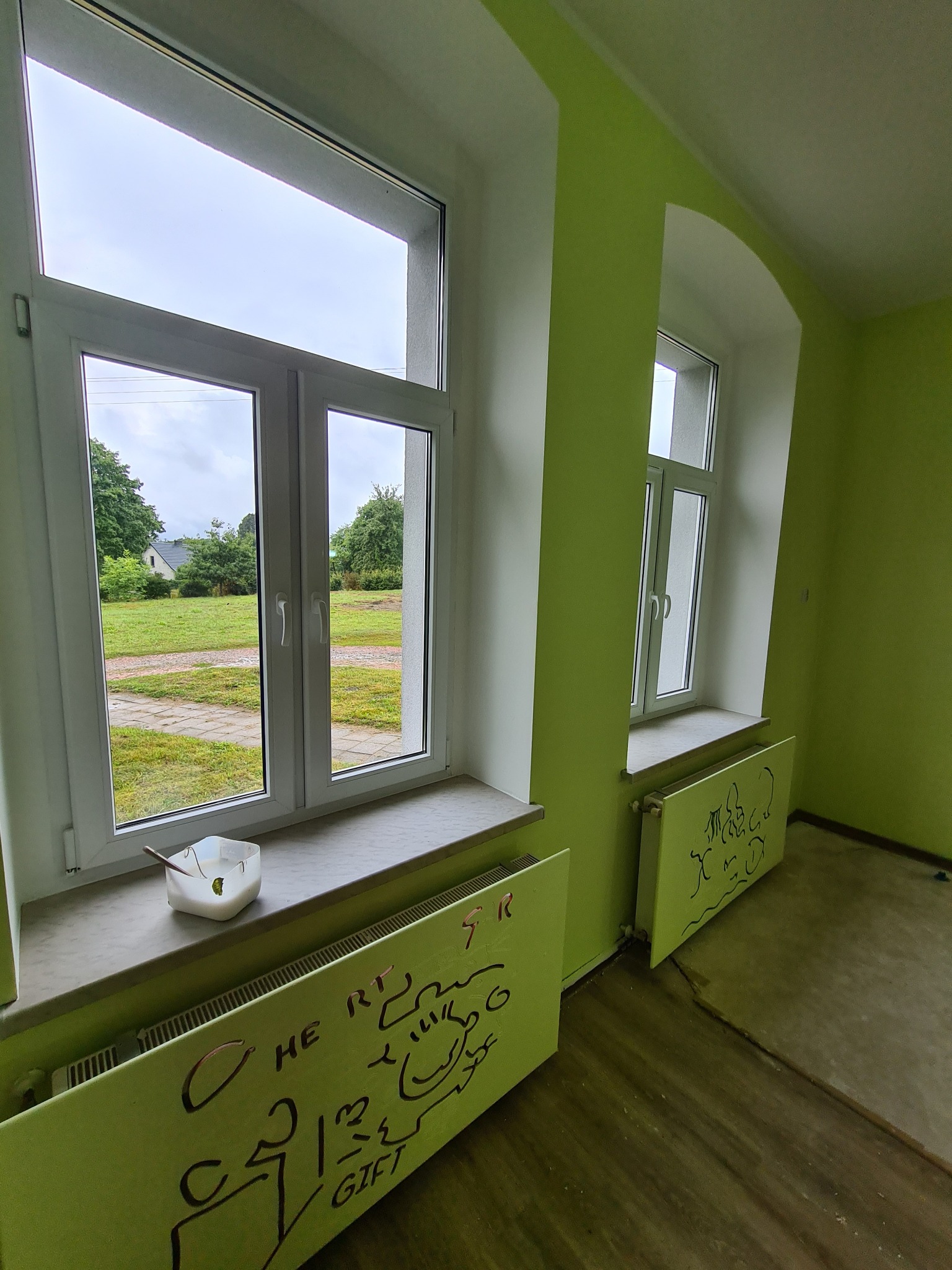 Pokój z zielonymi ścianami w trakcie malowania, widoczne dwa okna, na parapecie pojemnik z farbą, grzejniki z dziecięcymi rysunkami, podłoga częściowo wykończona.