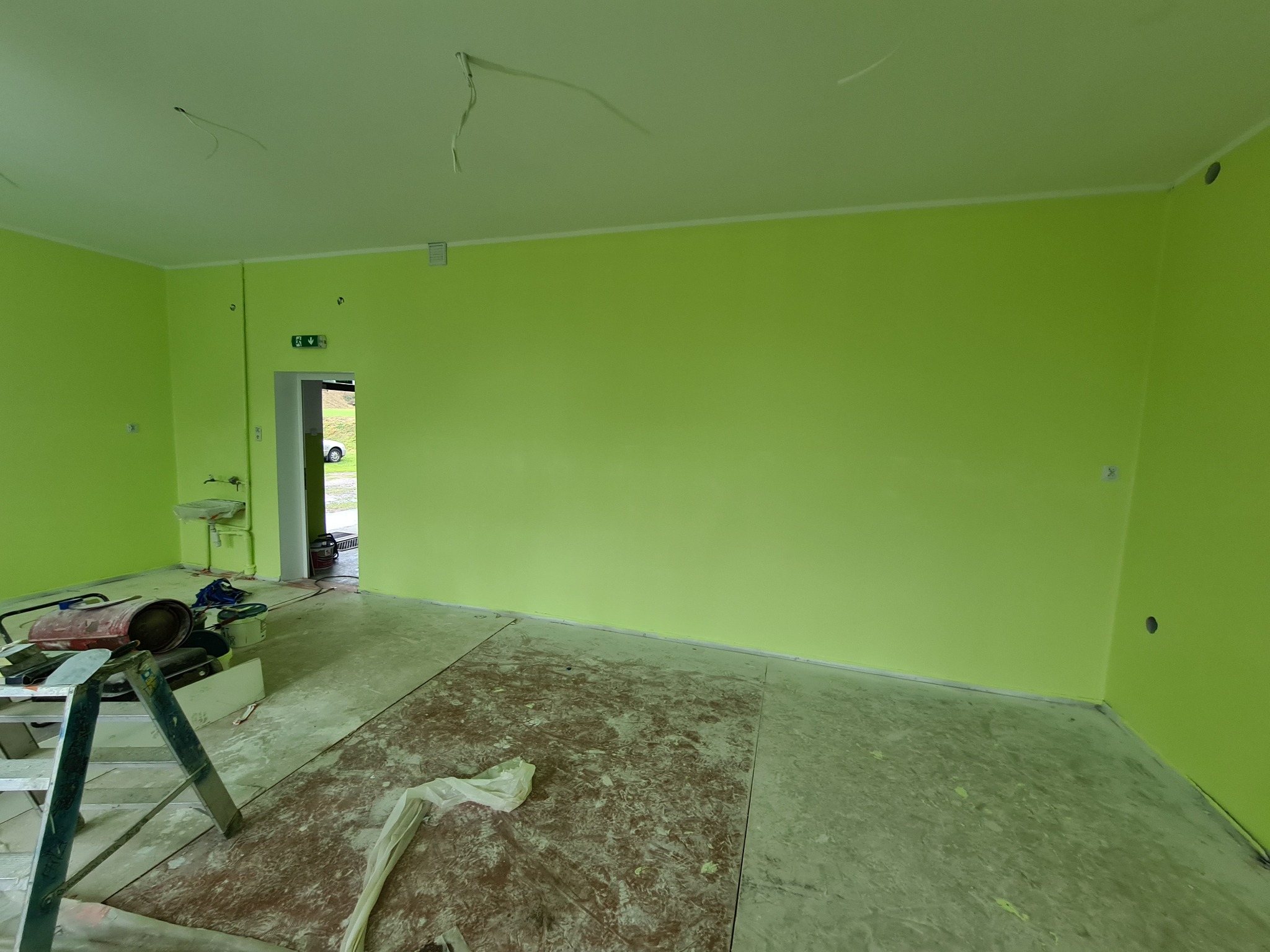 Pomalowane na jasnozielono wnętrze z widocznymi narzędziami malarskimi, drabina i zabezpieczona podłoga przed malowaniem, widoczne niedociągnięcia w postaci wystających kabli i niezabezpieczonych...