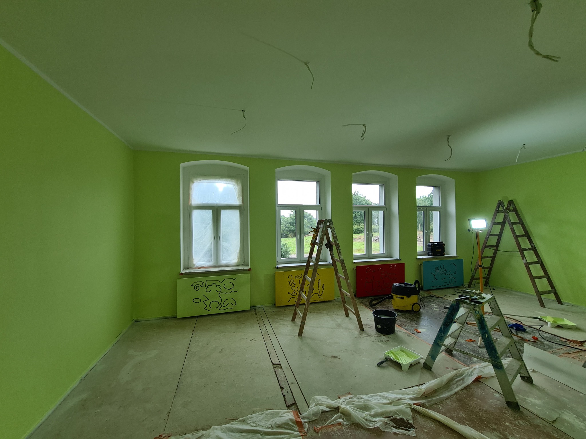 Wnętrze pokoju w trakcie malowania na zielono, widoczne trzy okna z częściowo zasłoniętymi szybami, grzejniki z dziecięcymi rysunkami, drabiny, lampa oświetlająca i narzędzia malarskie na podłodze.