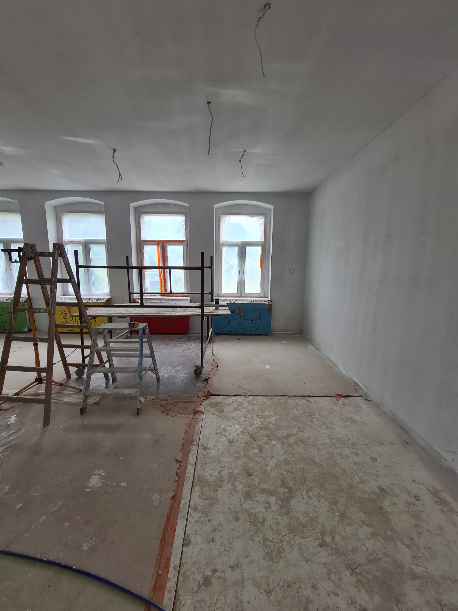 Pokój w trakcie prac wykończeniowych, ściany po nałożeniu gładzi, widoczne rusztowania, drabiny i zabezpieczone okna.