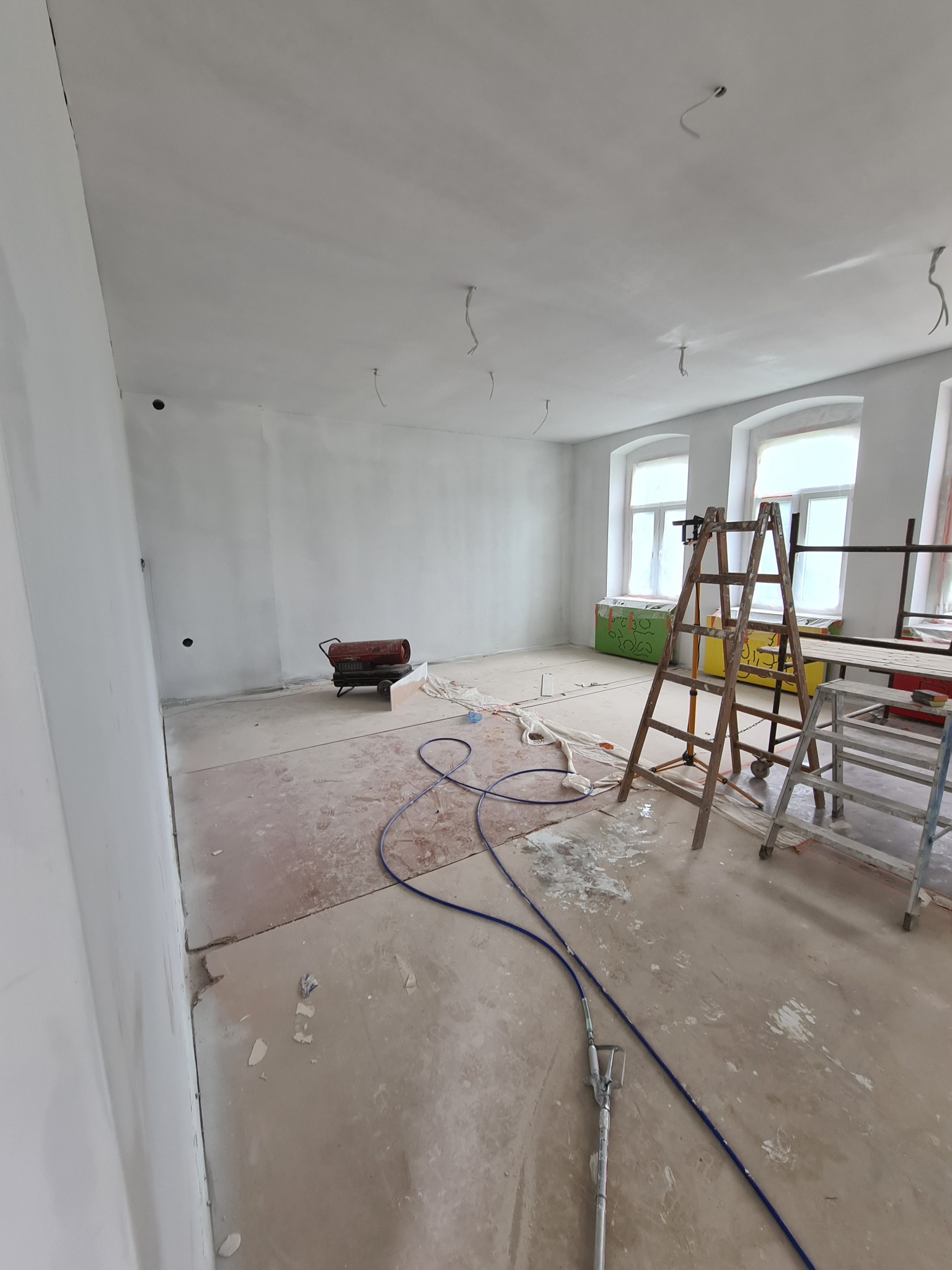 Wnętrze w trakcie prac przygotowawczych: ściany zagruntowane, widoczne drabiny, nagrzewnica, przewody i ślady po szpachlowaniu na podłodze, przygotowanie do malowania.
