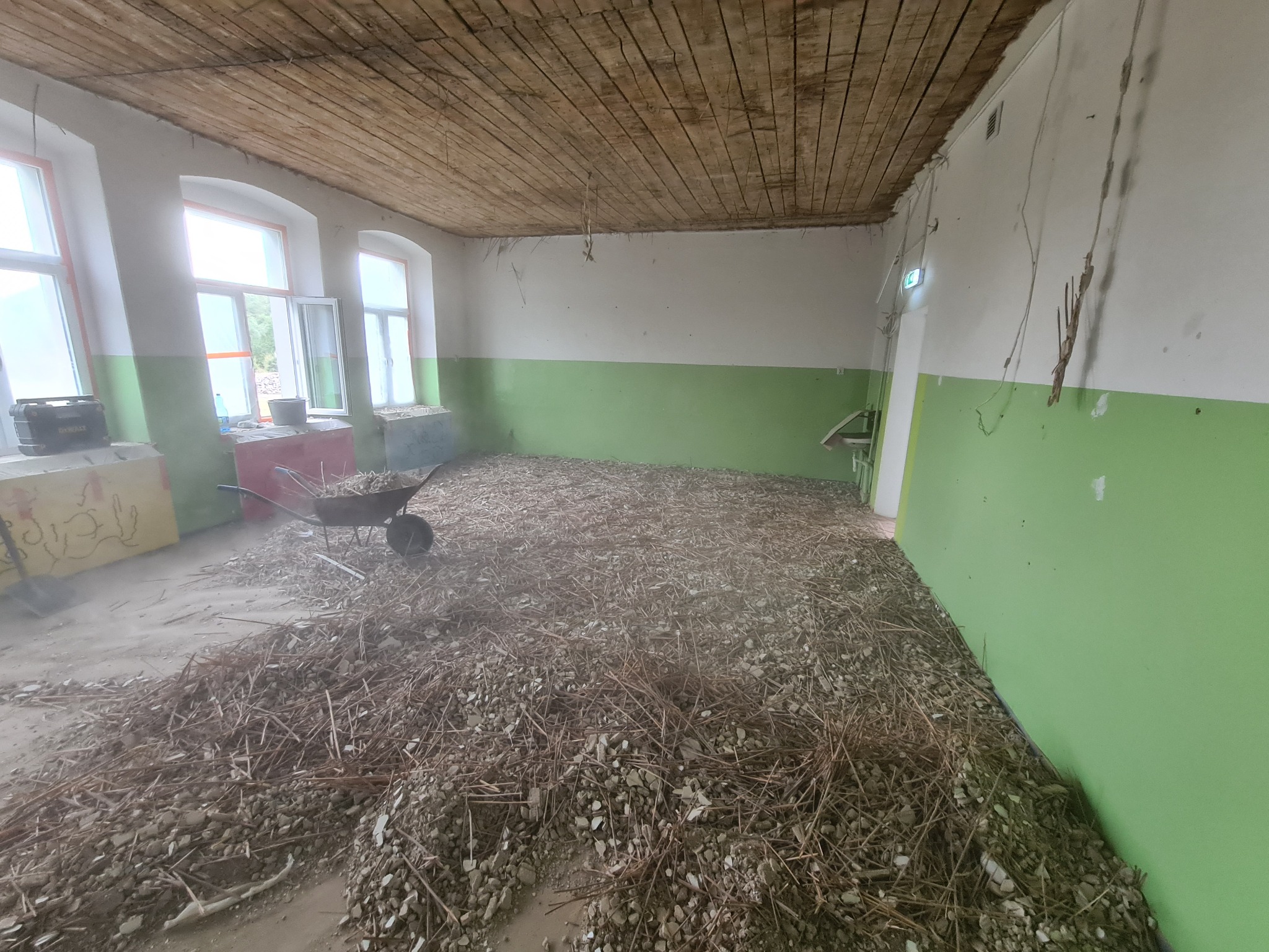 Pokój w trakcie demontażu: gruz i fragmenty drewna na podłodze, taczka w rogu, ściany pomalowane do połowy na zielono, drewniany sufit, okna z białymi ramami.