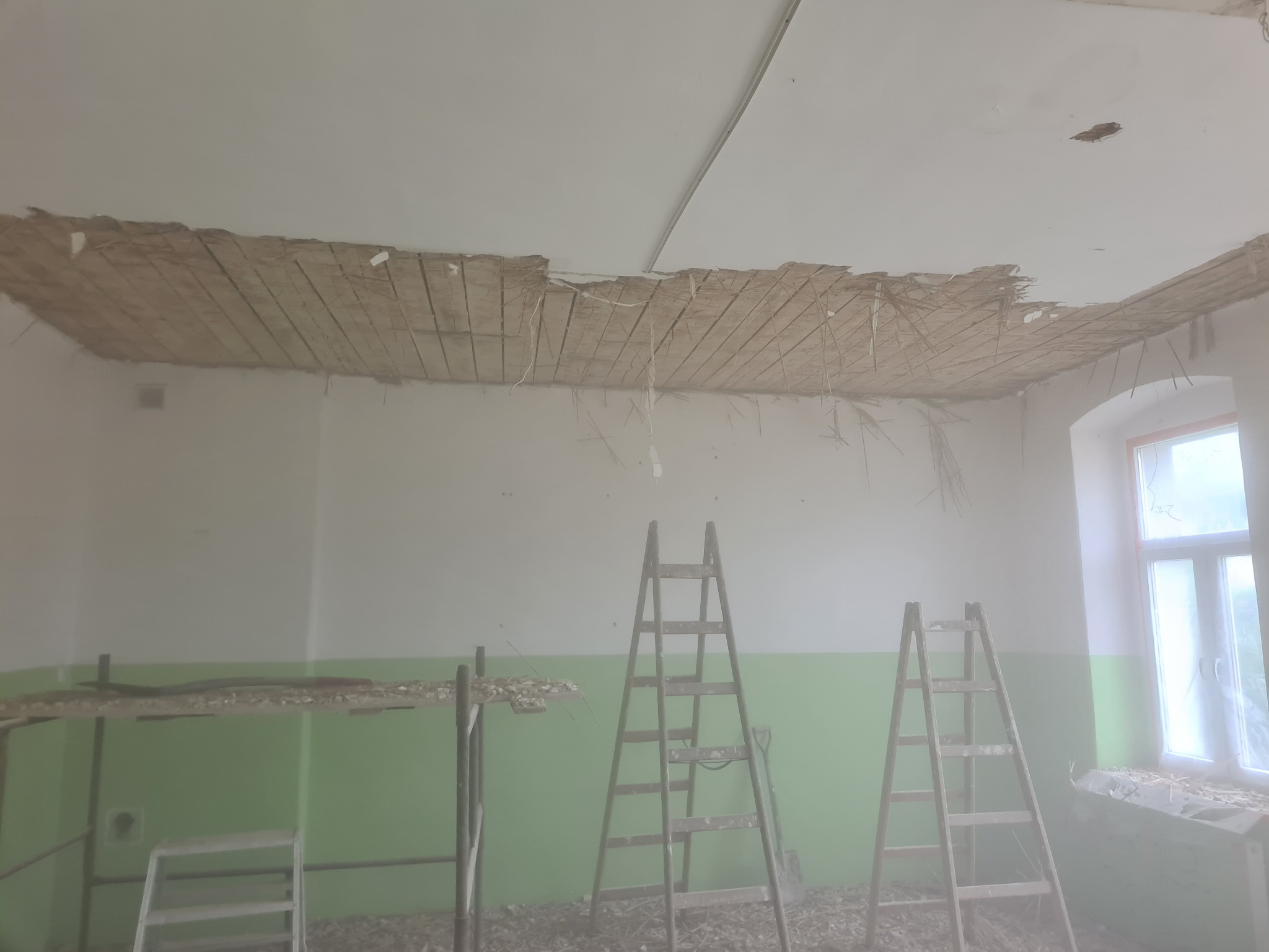 Demontaż starego sufitu w pomieszczeniu z zielonym pasem na ścianie; widoczne drewniane listwy i fragmenty tynku, dwie drabiny ustawione w pokoju, okno z parapetem również pokrytym resztkami...
