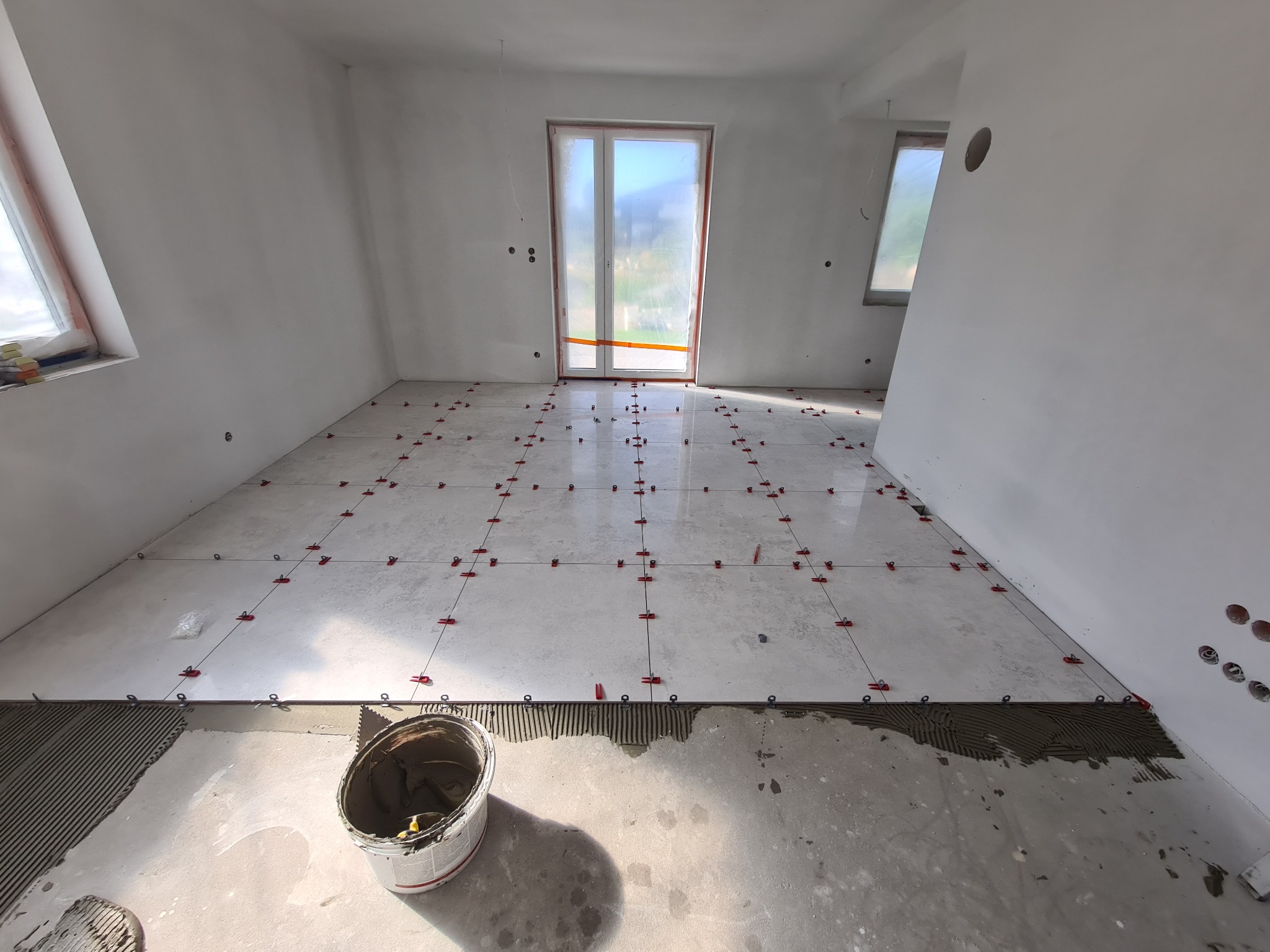 Jasnoszare płytki podłogowe w trakcie układania, z czerwonymi klipsami poziomującymi, widoczny fragment nałożonego kleju, białe ściany w surowym stanie, wiadro z zaprawą w rogu.