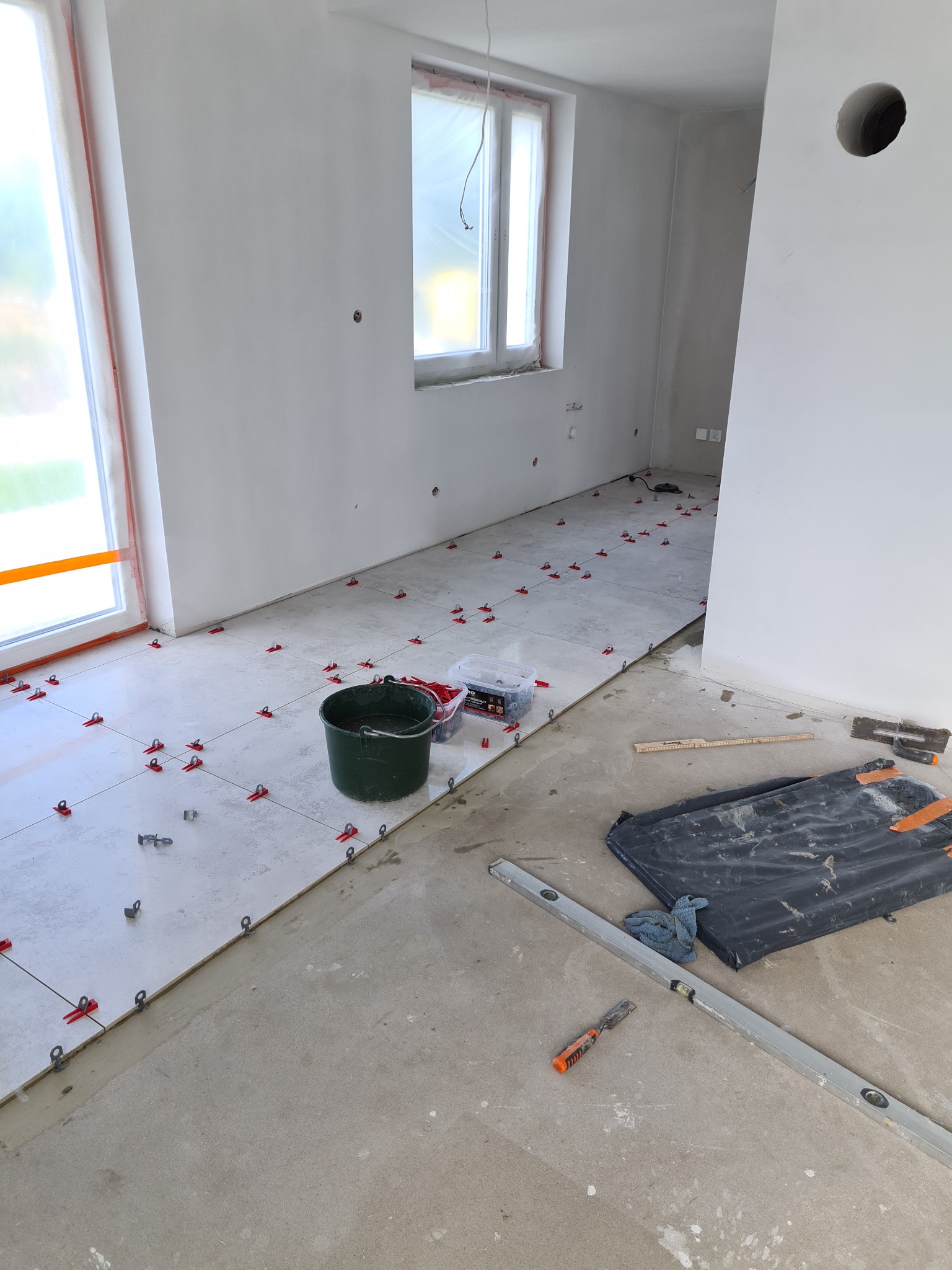 Układanie jasnych płytek podłogowych w pomieszczeniu, widoczne krzyżyki dystansowe, poziomica, wiadro i narzędzia glazurnicze.