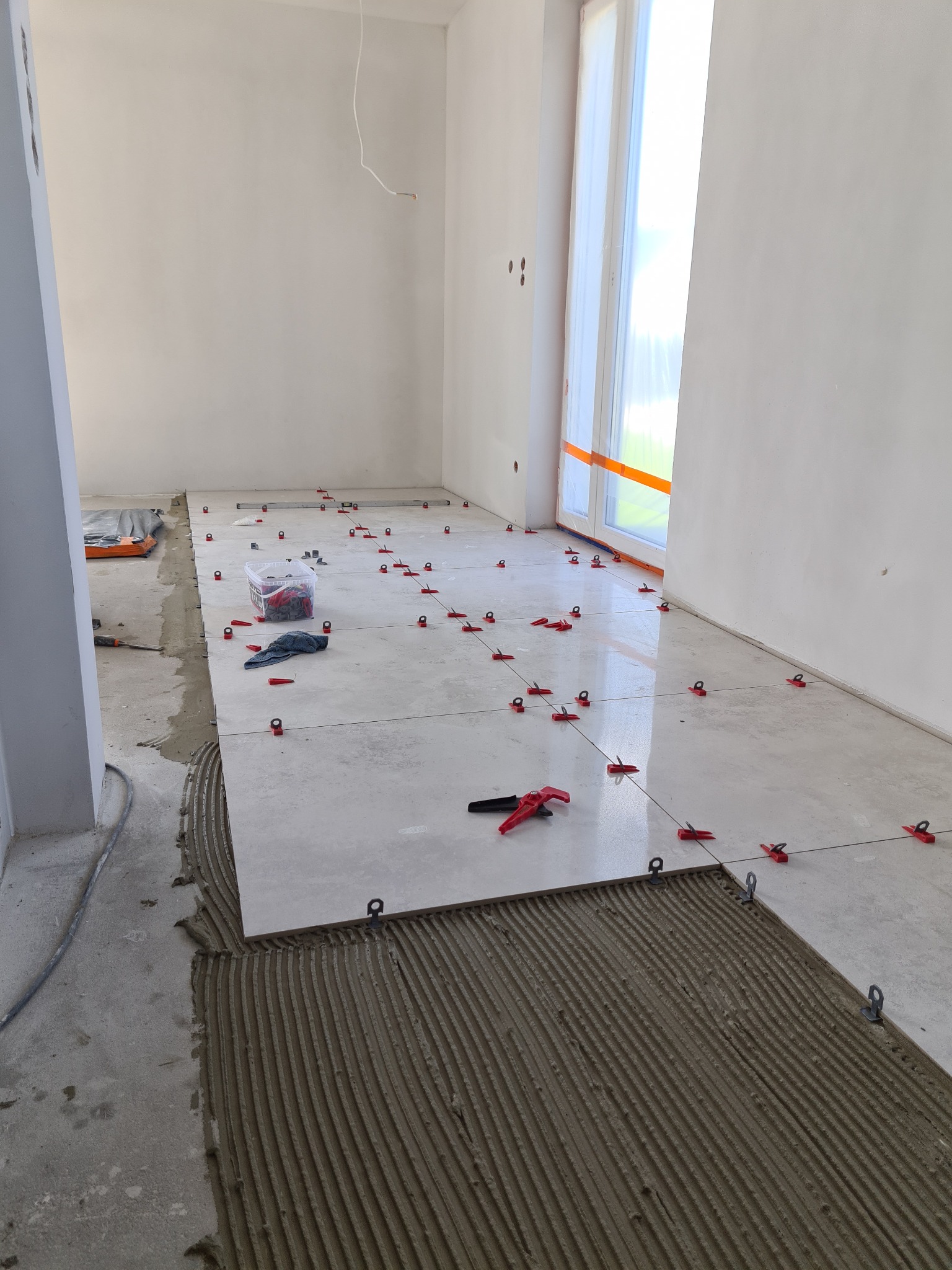 Układanie jasnych płytek podłogowych z użyciem systemu poziomowania, widoczny klej z charakterystycznymi rowkami, czerwone klipsy i kliny, w tle okno zabezpieczone folią.