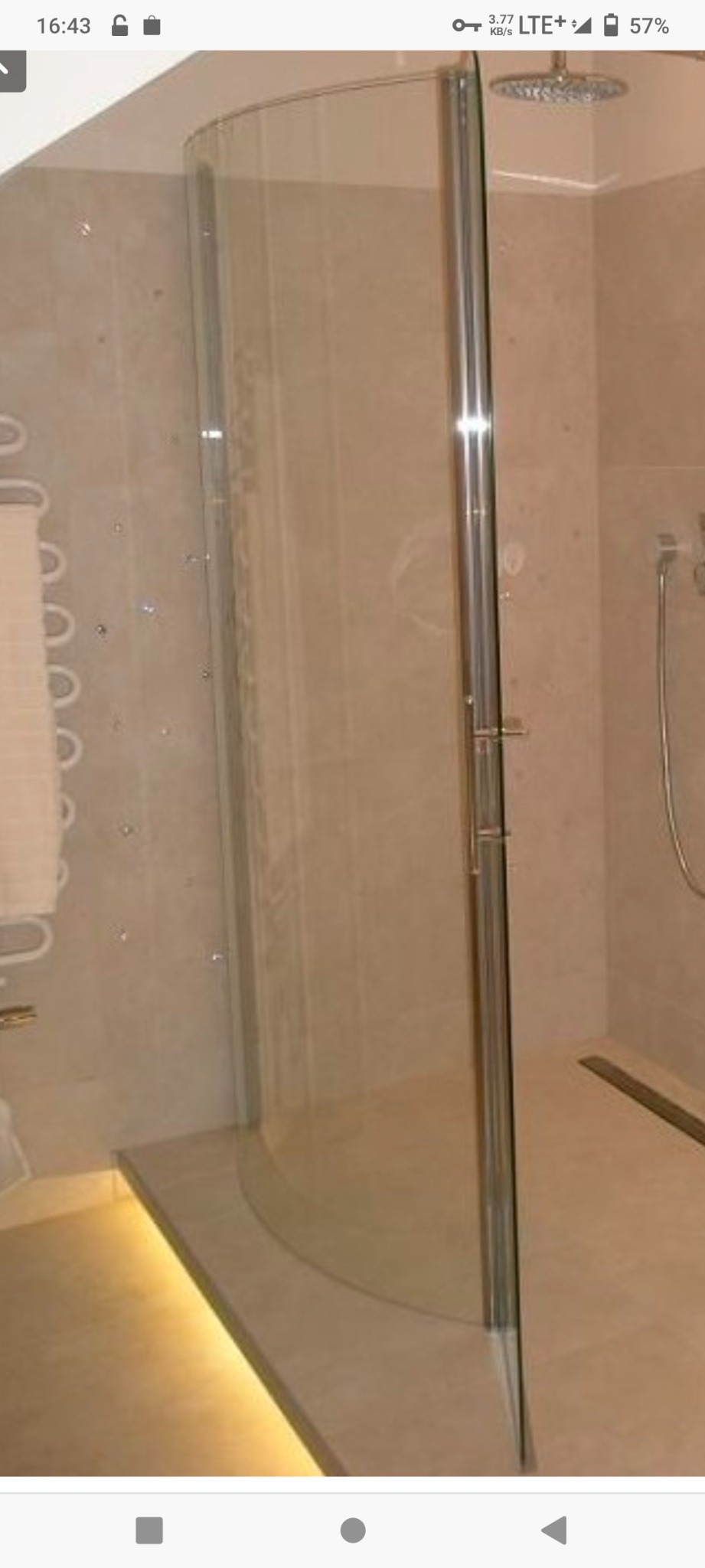 Zaokrąglona kabina prysznicowa ze szklanymi drzwiami i chromowanymi okuciami w jasnej łazience z beżowymi płytkami i ledowym oświetleniem pod podestem.