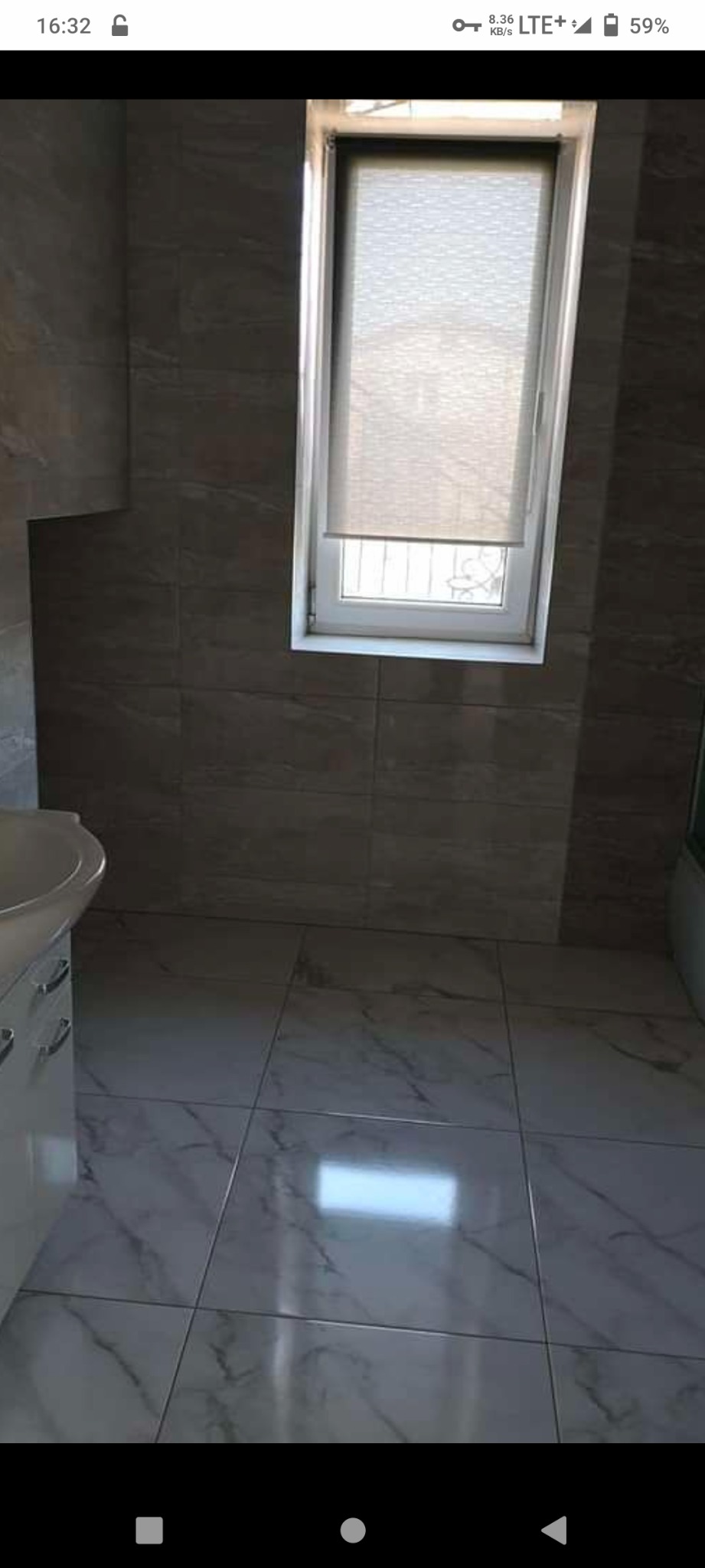 Wykończona łazienka z szarymi płytkami imitującymi kamień na ścianach i podłodze, biała szafka pod umywalką z chromowanymi uchwytami, okno z roletą w białej ramie.