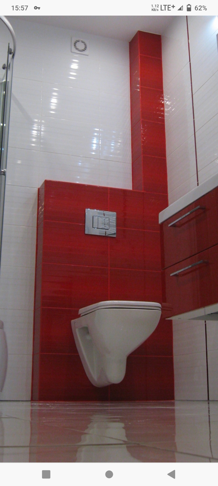 Ściana z czerwonych płytek w łazience z podwieszaną toaletą i chromowanym przyciskiem spłukującym, wentylator na białej ścianie, połyskująca podłoga.