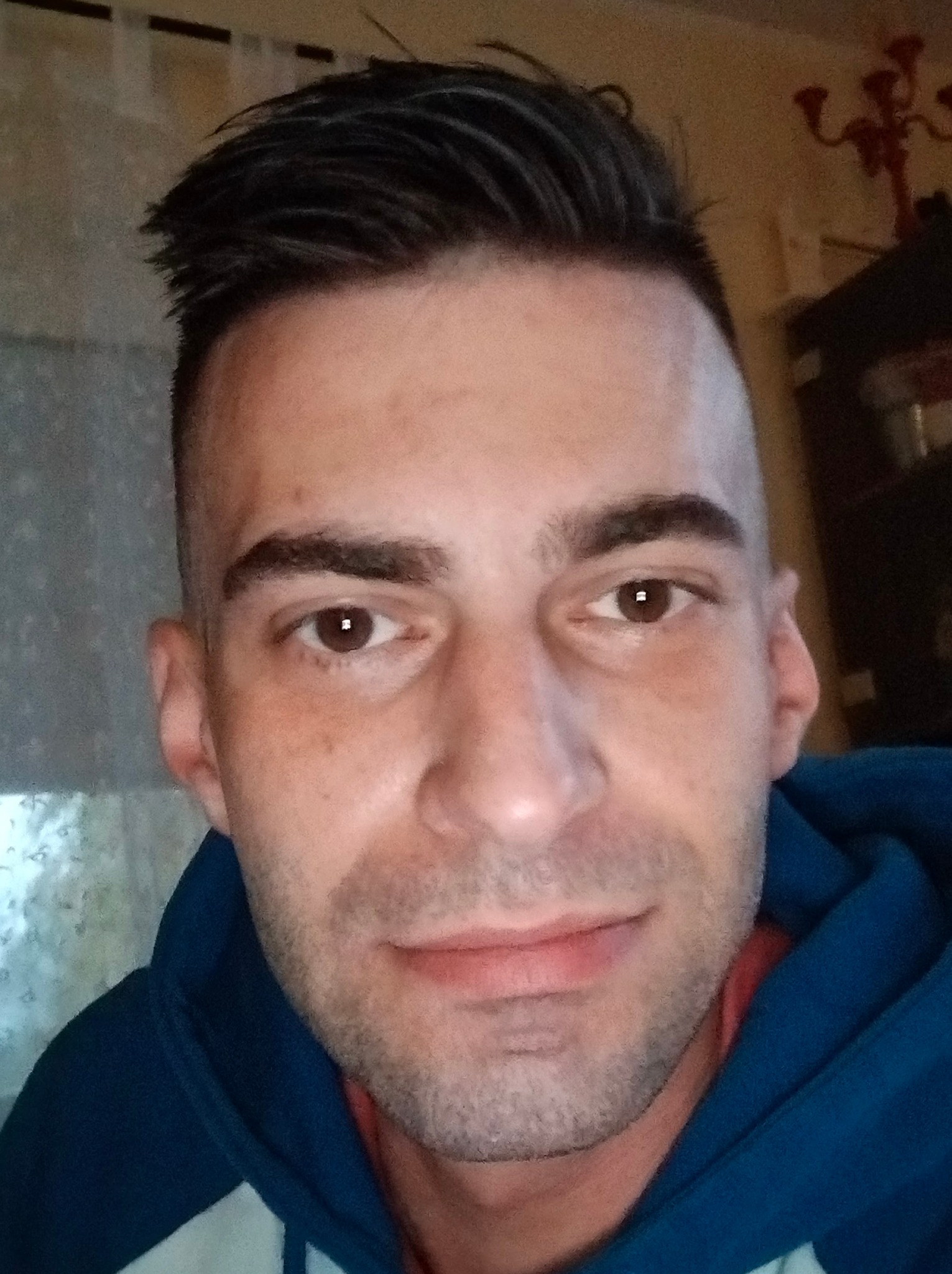 Portret mężczyzny z krótkimi, ciemnymi włosami i brązowymi oczami, ubrany w niebieską bluzę z kapturem, na rozmytym tle wnętrza.