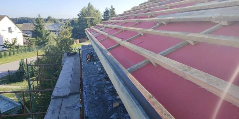 Montaż izolacji dachu z czerwonej membrany i drewnianych łat, widok z rusztowania na tle zieleni i zabudowań.