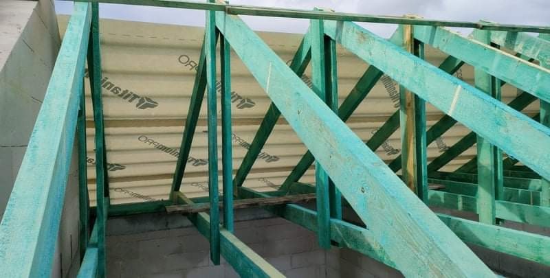 Konstrukcja dachu z impregnowanego drewna w kolorze turkusowym, widoczne płyty izolacyjne z nadrukiem logo producenta, ściany z bloczków betonowych w tle.