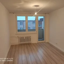 Puste, jasne pomieszczenie po remoncie z panelami podłogowymi imitującymi drewno, białymi ścianami, grzejnikiem i oknem balkonowym z widokiem na blok.