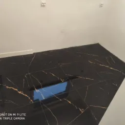 Ukończona podłoga w łazience wyłożona czarnymi płytkami z złotymi żyłkami, widoczny biały grzejnik i gniazdka elektryczne na białej ścianie.