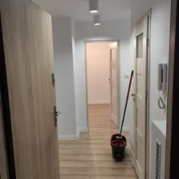 Korytarz w mieszkaniu po remoncie, z panelami podłogowymi w odcieniach drewna, świeżo umytymi mopem z czerwonym wiadrem, widoczne białe ściany i sufit z trzema okrągłymi lampami, oraz dwie...