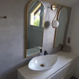 Nowoczesna łazienka z owalną umywalką nablatową, czarną armaturą i dwoma lustrami w złotych ramach, oświetlona ozdobną lampą ścienną.