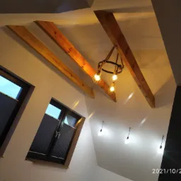 Poddasze z drewnianymi belkami stropowymi, oświetleniem w stylu industrialnym i oknami z żaluzjami plisowanymi.