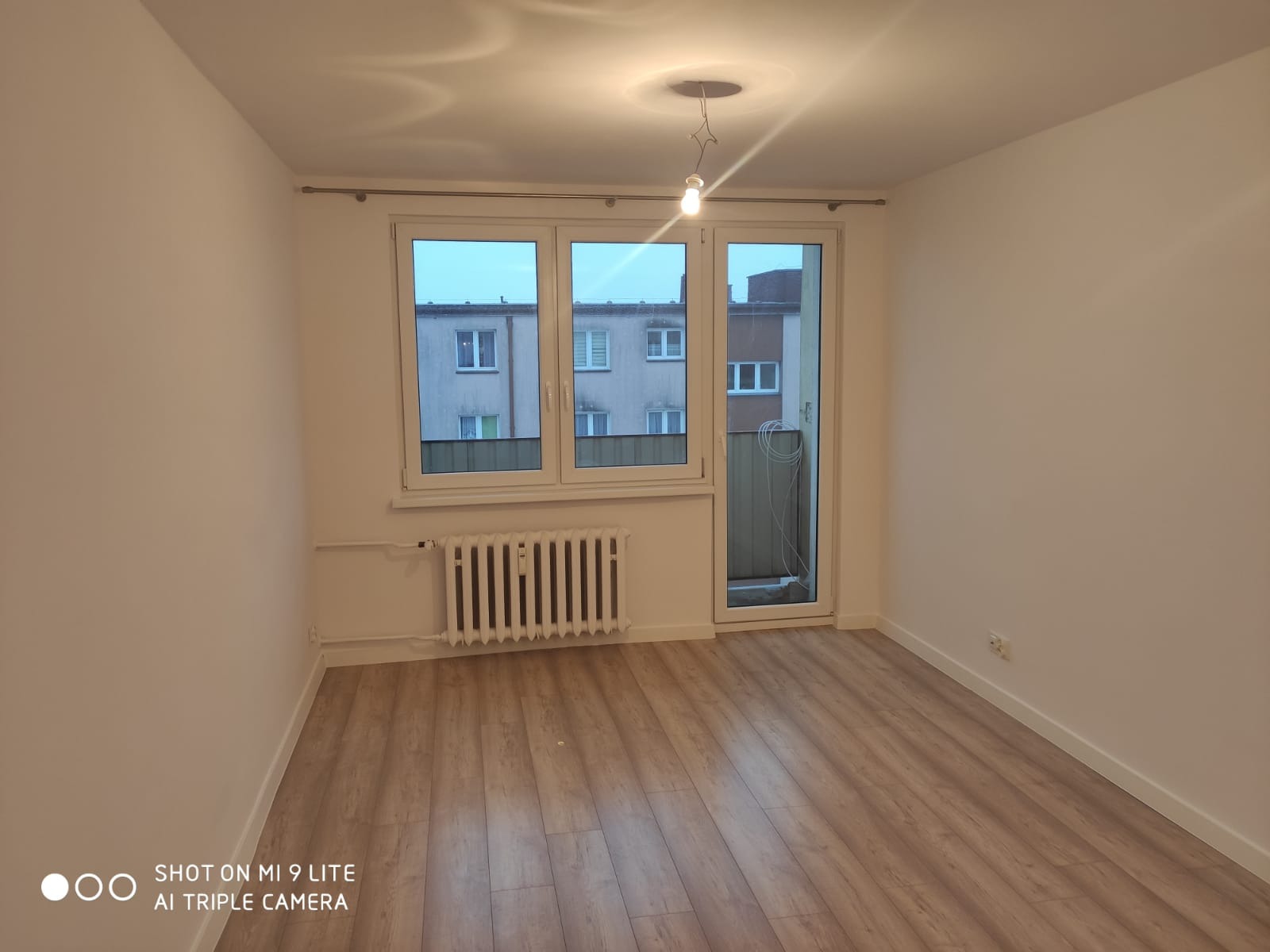 Puste, jasne pomieszczenie po remoncie z panelami podłogowymi imitującymi drewno, białymi ścianami, grzejnikiem i oknem balkonowym z widokiem na blok.