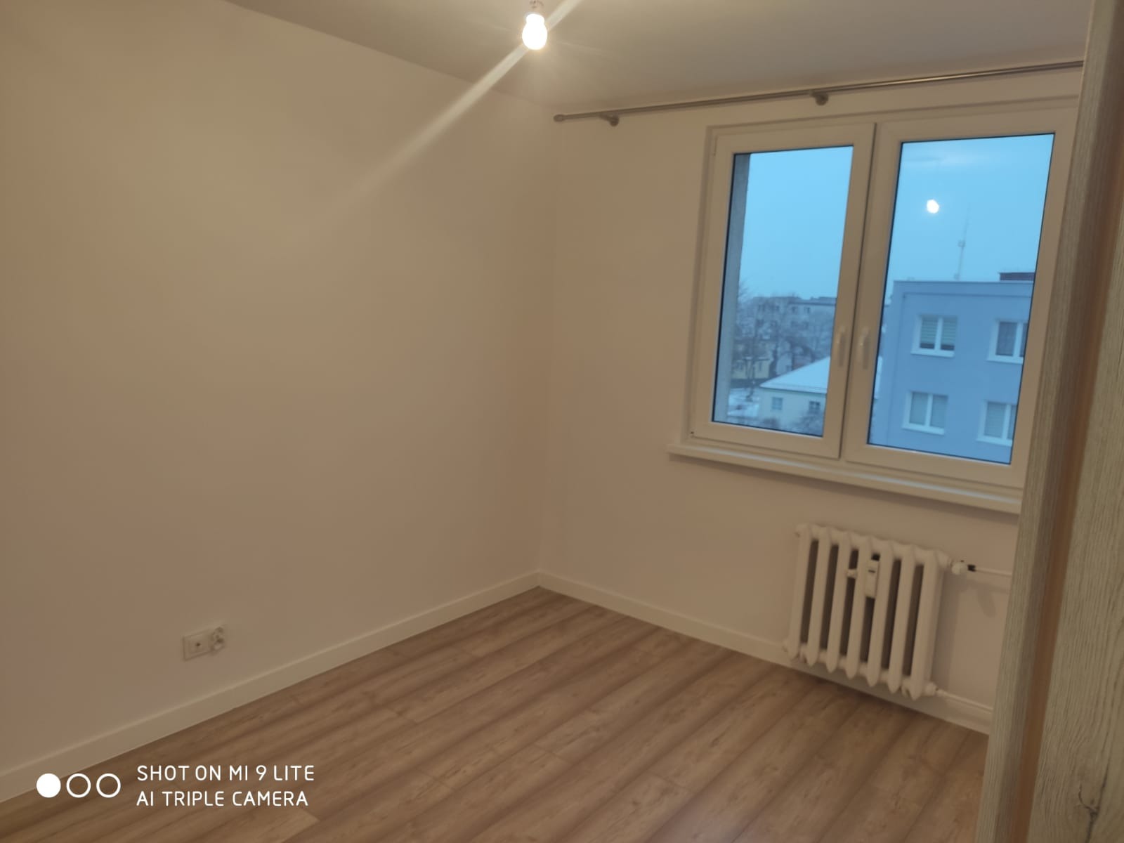 Pusty pokój po remoncie: jasne ściany, drewnopodobne panele podłogowe, białe okno z widokiem na budynek i zimowe drzewa, biały grzejnik pod oknem.