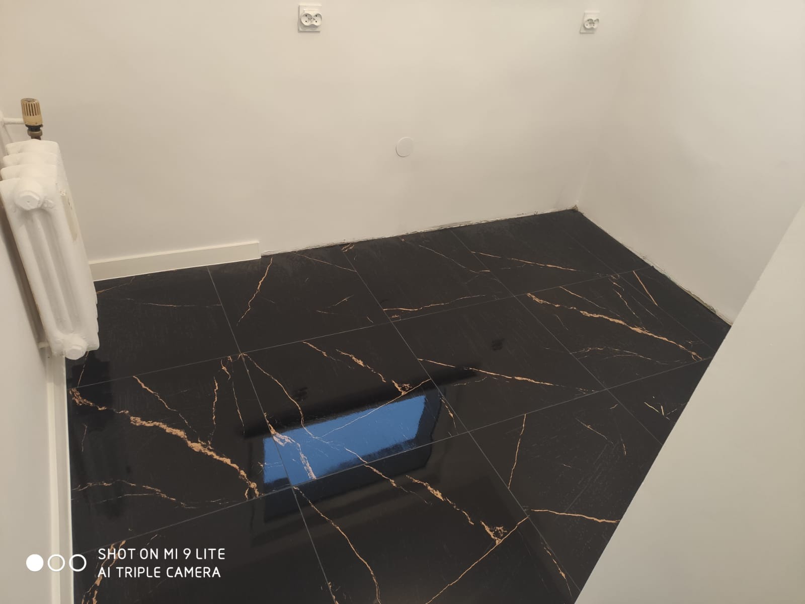 Ukończona podłoga w łazience wyłożona czarnymi płytkami z złotymi żyłkami, widoczny biały grzejnik i gniazdka elektryczne na białej ścianie.