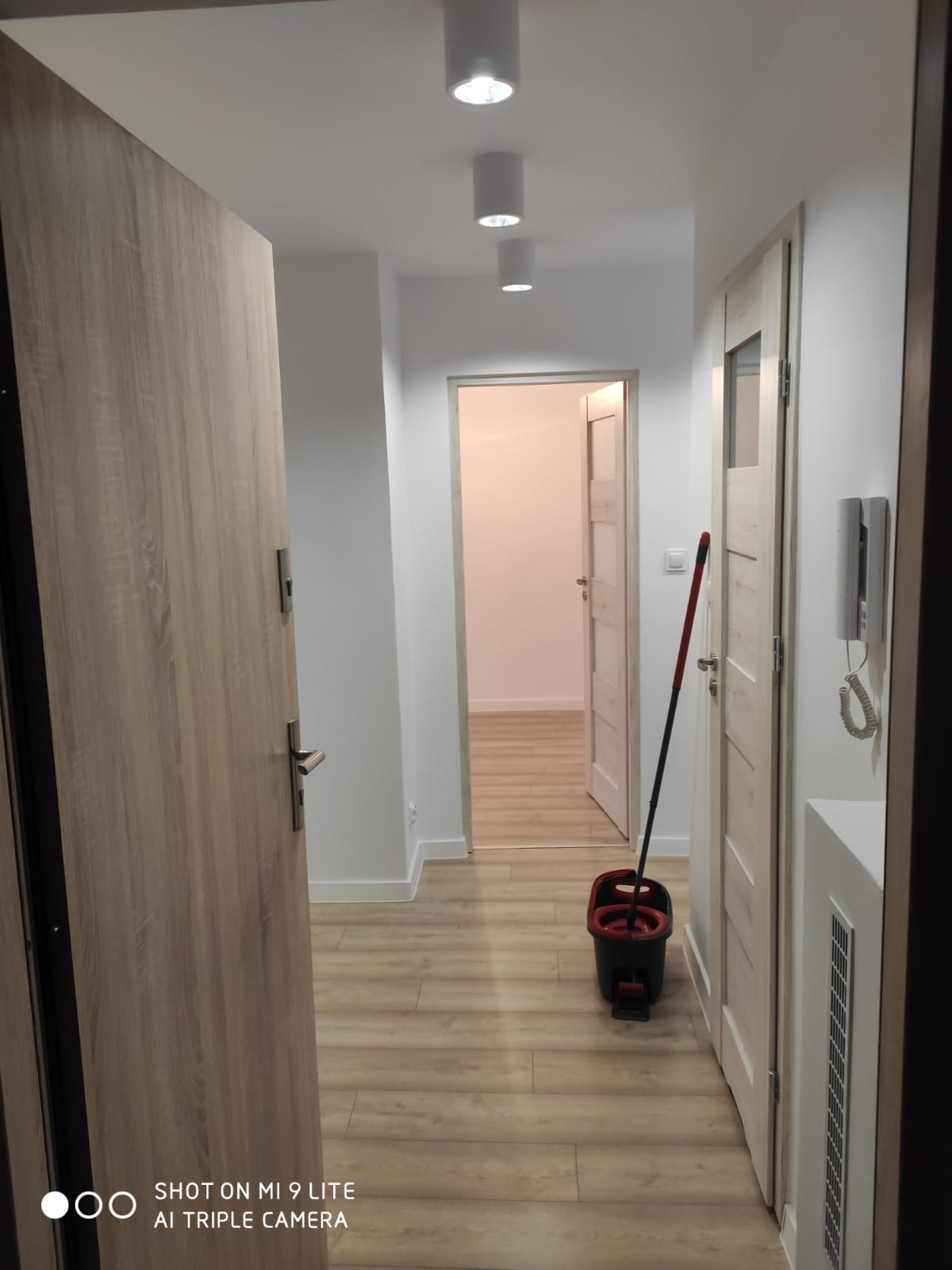 Korytarz w mieszkaniu po remoncie, z panelami podłogowymi w odcieniach drewna, świeżo umytymi mopem z czerwonym wiadrem, widoczne białe ściany i sufit z trzema okrągłymi lampami, oraz dwie...