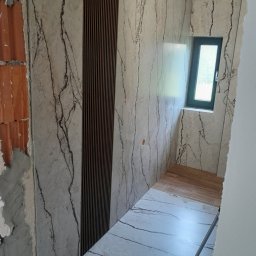 uslugi remontowo budowlane - Wnętrze łazienki w trakcie remontu: ściany wyłożone płytkami imitującymi marmur z brązowymi żyłami, dekoracyjny panel z pionowych drewnianych listew, fragment odsłoniętej cegły, małe okno z ciemną...