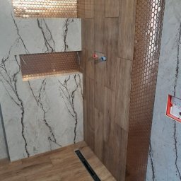 uslugi remontowo budowlane - Nowoczesna aranżacja łazienki w Grywałdzie: połączenie płytek imitujących drewno, marmuru i mozaiki heksagonalnej, z podświetlanymi wnękami i odpływem liniowym w podłodze.