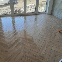 uslugi remontowo budowlane - Świeżo ułożona, błyszcząca podłoga z płytek imitujących drewno ułożonych w jodełkę, widok na okno z widokiem na plac budowy i fragment drabiny malarskiej.