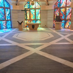 uslugi remontowo budowlane - Wnętrze kaplicy z geometryczną posadzką w odcieniach szarości i beżu, marmurowe ściany oraz witraże w oknach, rzucające kolorowe refleksy świetlne na podłogę.