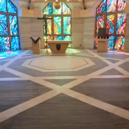 Wnętrze kaplicy z geometryczną posadzką w odcieniach szarości i beżu, marmurowe ściany oraz witraże w oknach, rzucające kolorowe refleksy świetlne na podłogę.