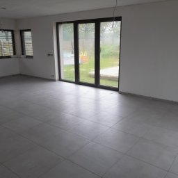 uslugi remontowo budowlane - Wnętrze pustego pokoju z szarymi, kwadratowymi płytkami podłogowymi, dużym oknem balkonowym z widokiem na ogród z placem zabaw i dwoma mniejszymi oknami z żaluzjami, ściany pomalowane na biało...