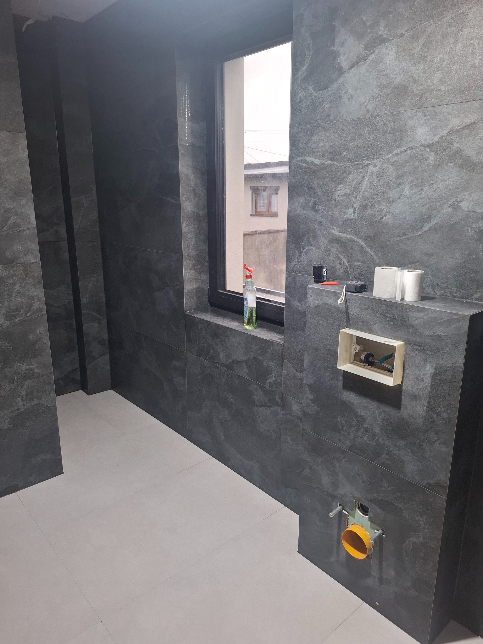 Ciemne, marmurkowe płytki ścienne w łazience w trakcie montażu, widoczne przyłącza do toalety, okno z widokiem na sąsiedni budynek, butelka środka czyszczącego na parapecie, częściowo wykończona...